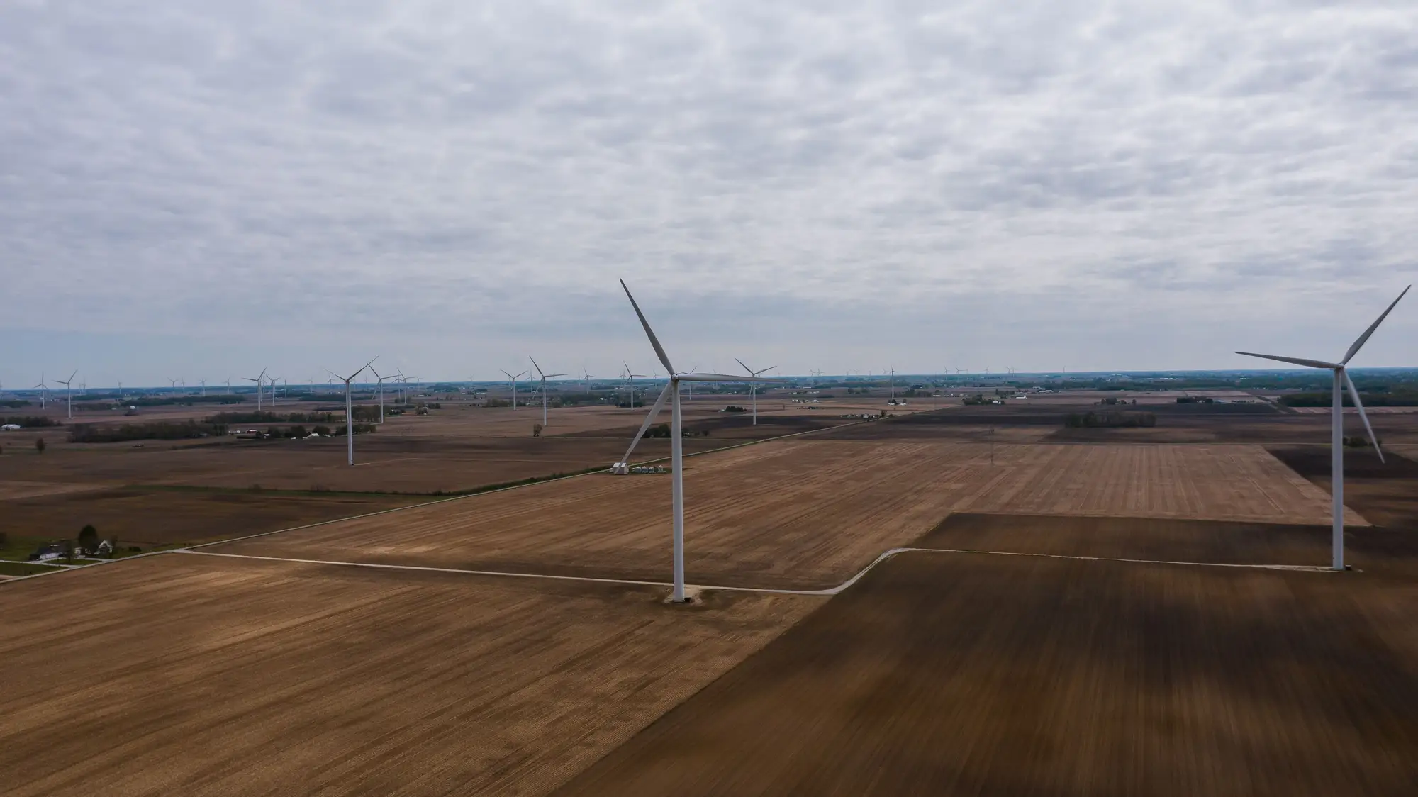A string of wind turbines on flat ploughed farmland under a grey sky, seen from a drone